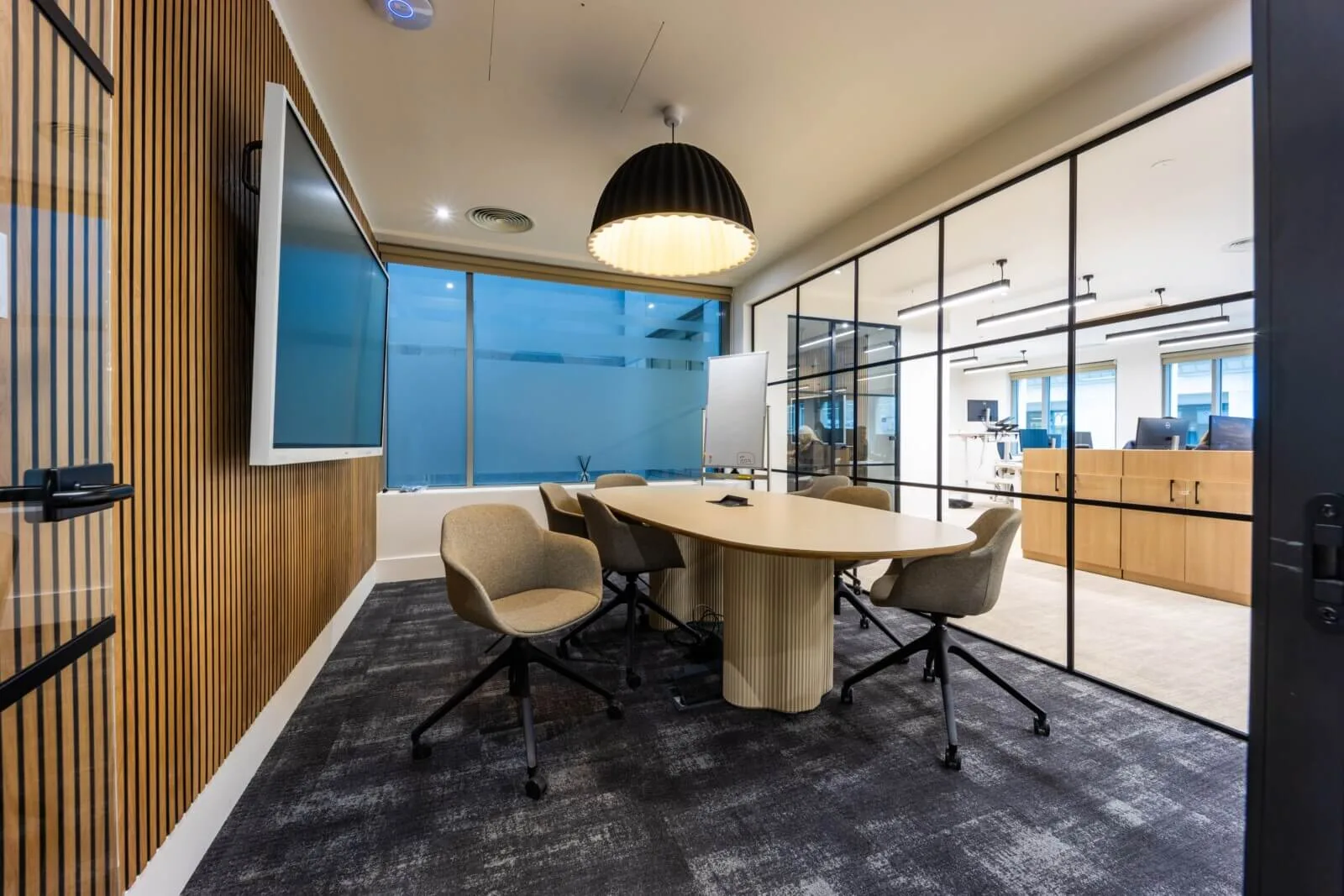 Glazed meeting room with an oval oak table, pendant light and adjacent open workspace