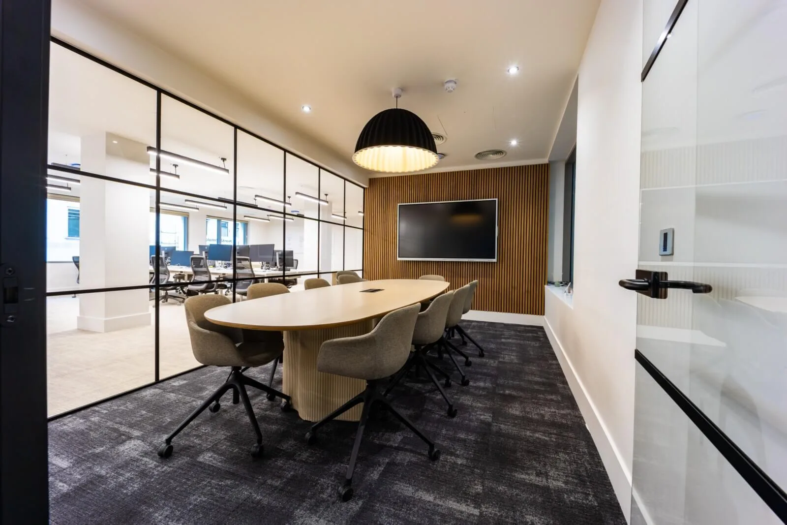 Oval oak meeting table with upholstered chairs, a slatted media wall and black-framed glazing