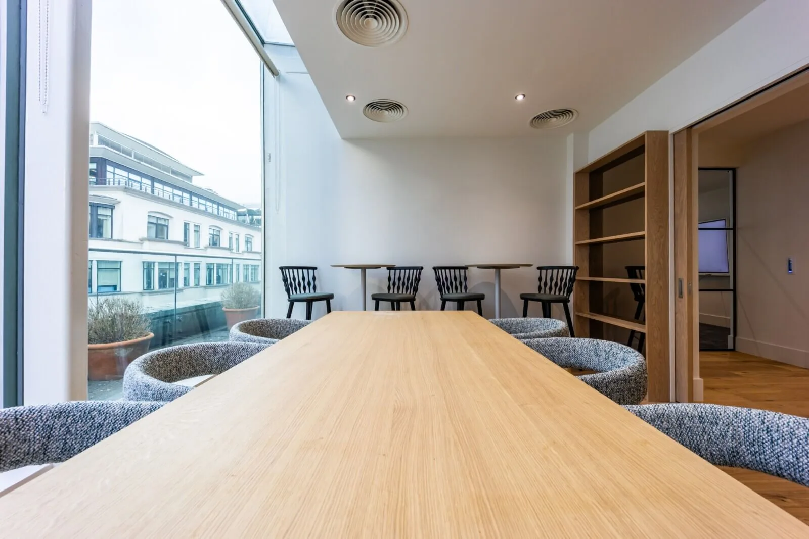 Long pale oak project table beside full-height windows and cafe-height seating