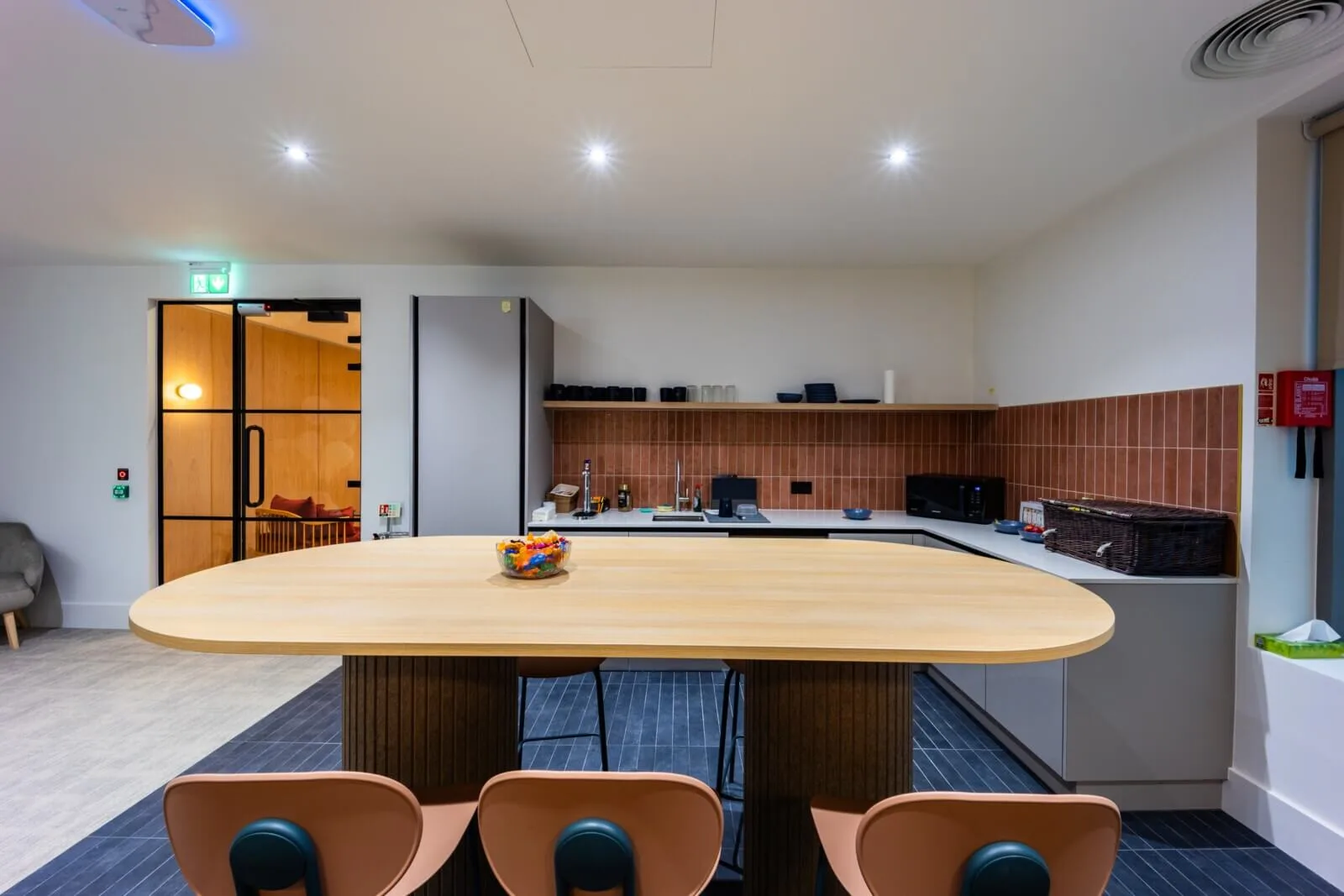 Terracotta-tiled kitchen with a long pale oak island and muted salmon chairs